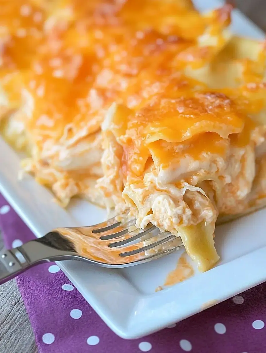 A fork is lifting a portion of cheesy, creamy chicken lasagna from a white plate, with a purple dotted napkin underneath.